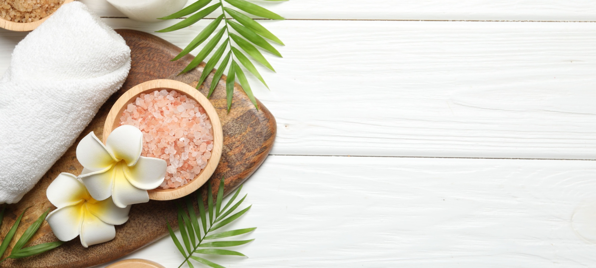 Flachgelegtes Wellness-Arrangement auf wei&szlig;em Holz mit Badesalz, Handtuch, Massageb&uuml;rste, Kerze und tropischen Bl&auml;ttern sowie Frangipani-Bl&uuml;ten auf der linken Seite.