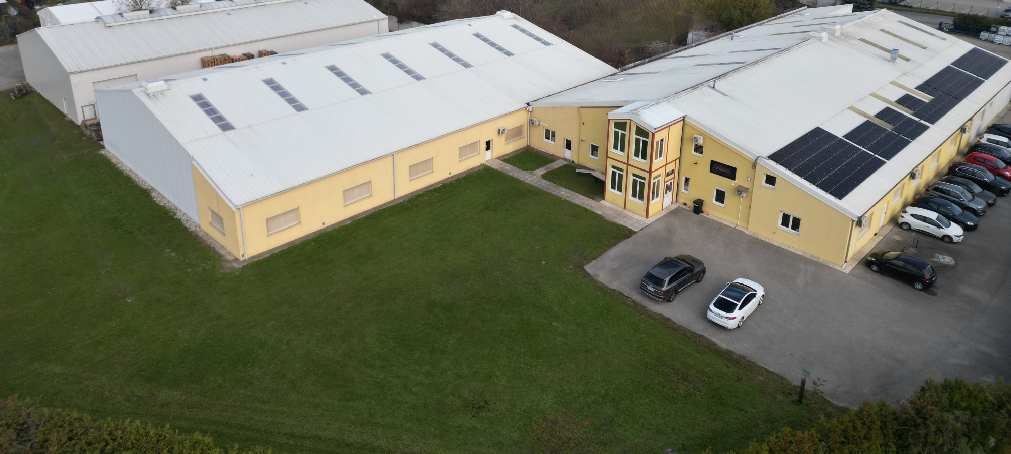 Luftaufnahme eines gro&szlig;en, L-f&ouml;rmigen Gewerbegeb&auml;udes mit gelber Fassade, wei&szlig;en D&auml;chern, Solaranlagen und einem angrenzenden Parkplatz mit mehreren Autos.