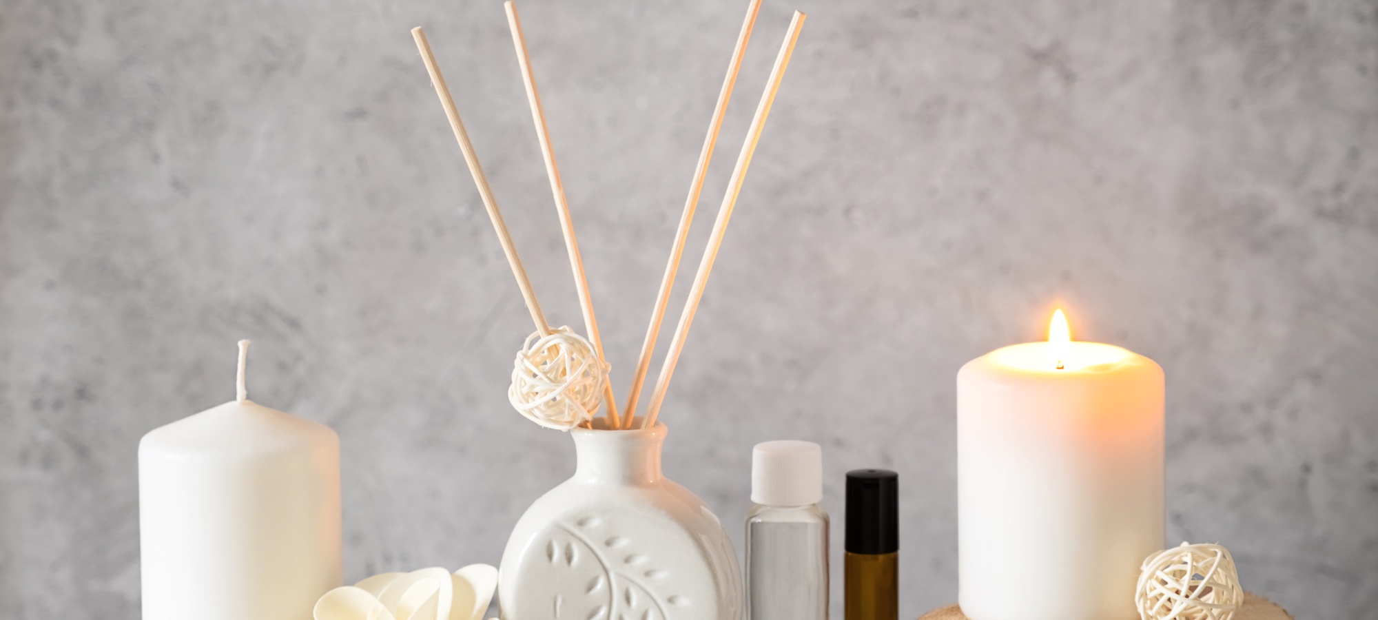 Ein wei&szlig;er Aroma-Diffuser mit Holzst&auml;bchen und zwei 10ml Glasflaschen stehen zwischen zwei wei&szlig;en Stumpenkerzen, wovon eine auf einer Holzscheibe brennt.