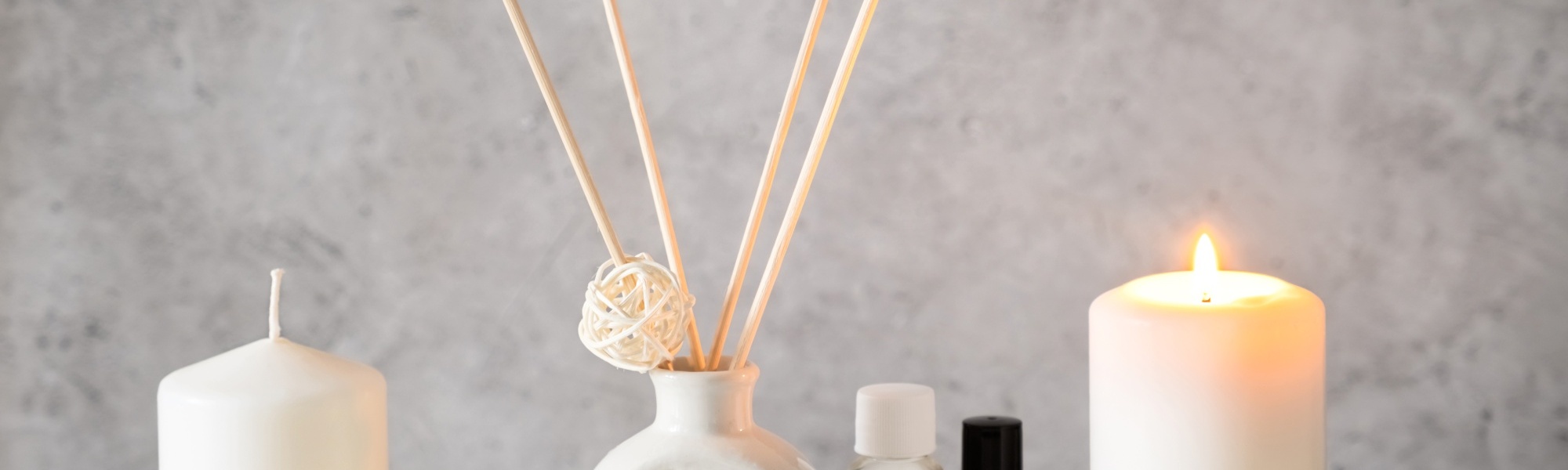 Ein wei&szlig;er Aroma-Diffuser mit Holzst&auml;bchen und zwei 10ml Glasflaschen stehen zwischen zwei wei&szlig;en Stumpenkerzen, wovon eine auf einer Holzscheibe brennt.