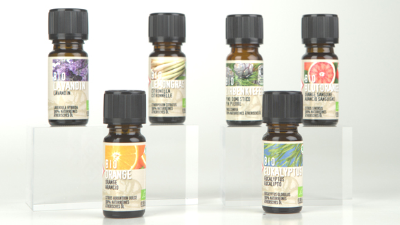 Sechs kleine, braune Fl&auml;schchen mit Bio-Aroma&ouml;len stehen auf transparenten Podesten vor einem hellen Hintergrund.