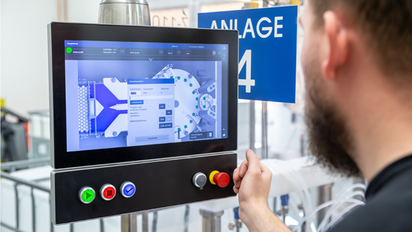 Das Bild zeit eine Person die einen digitalen Bedienbildschirm einer Industrieanlage bedient, Produktionsdaten und Steuerungsmen&uuml;s anzeigt; darunter befindet sich ein Bedienfeld mit mehreren farbigen Tasten und einem Not-Aus-Schalter.