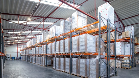 Gro&szlig;e, gut ausgeleuchtete Lagerhalle mit hohen Palettenregalen aus Metall, die dicht mit in Stretchfolie verpackten Kartons und Waren best&uuml;ckt sind.