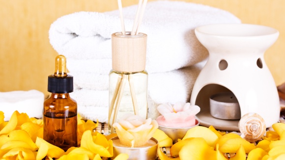 eine Szene mit brauner Glasflasche und Pipette, ein Aromasticksdiffuser mit Holzkappe, eine Keramikduftlampe in wei&szlig;, im Vordergrund gelbe Bl&uuml;tenbl&auml;tter und im Hintergrund zwei wei&szlig;e Handt&uuml;cher