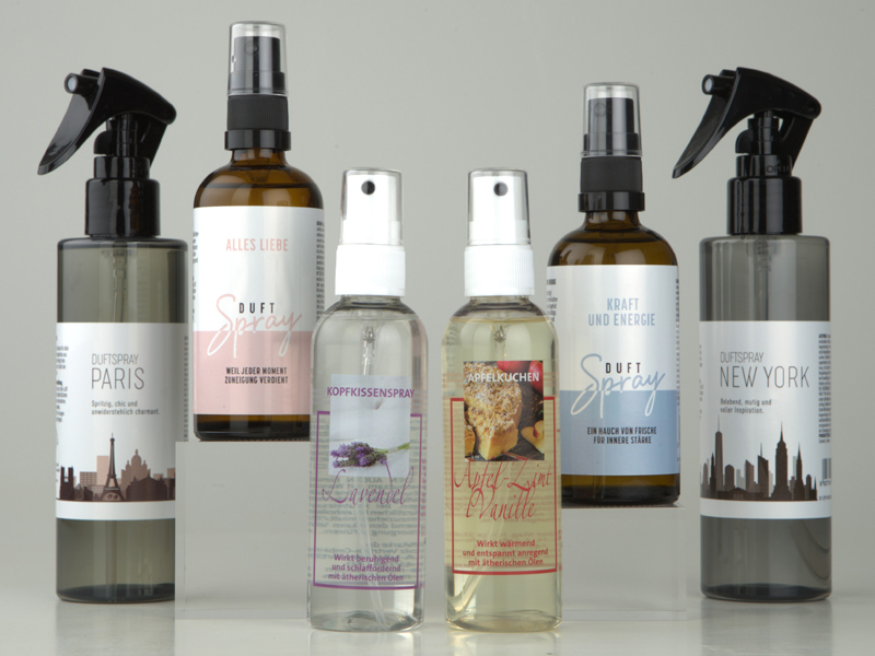 Neun transparente Spr&uuml;hflaschen mit verschiedenen Aroma- und Raumsprays stehen auf kleinen, klarsichtigen Podesten vor wei&szlig;em Hintergrund.