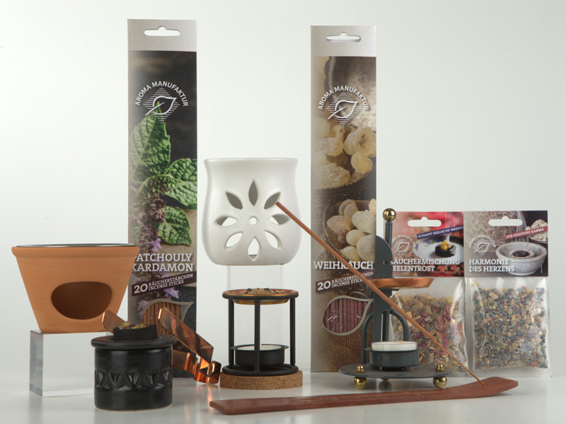 Verschiedene R&auml;ucherutensilien, darunter ein wei&szlig;es St&ouml;vchen, R&auml;ucherst&auml;bchen-Packungen, eine Halterung mit brennendem St&auml;bchen und kleine Beutel mit R&auml;uchermischungen vor wei&szlig;em Hintergrund.