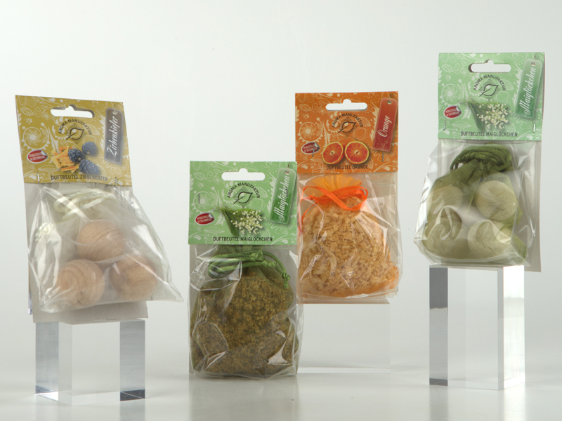 Vier verpackte Duftbeutel der Marke Aroma Manufaktur, gef&uuml;llt mit Holzkugeln oder Kr&auml;utern, stehen auf transparenten Podesten vor wei&szlig;em Hintergrund.